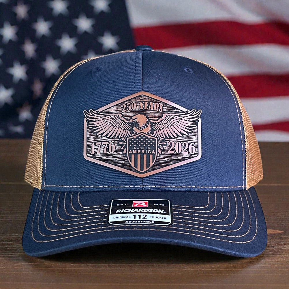 Blue trucker hat with eagle emblem and '250 Years of America' design on a wooden surface with an American flag in the background.