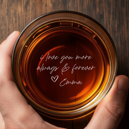 Hand holding a glass of amber liquid with 'i love you more always & forever Emma' text on a wooden surface.